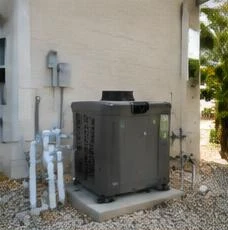 Heat pump pool heater hidden in a fenced side yard between a stucco wall and wooden fence